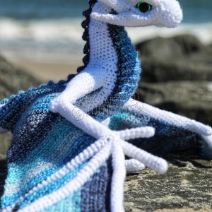 May include: A crocheted blue and white dragon with green eyes. The dragon is laying on a rock with the ocean in the background. The text "CRAFTY INTENTIONS" is on the image.  PDF.