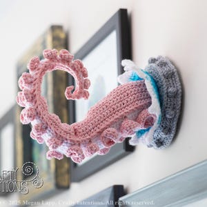 May include: A crocheted octopus tentacle wall hanging in shades of pink, white, and gray. The tentacle is curled and detailed, with a textured appearance. The piece is mounted on a wall, with a framed picture in the background. The overall theme is nautical decor.