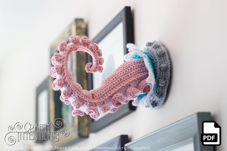May include: A crocheted octopus tentacle wall hanging in shades of pink, white, and gray. The tentacle is curled and detailed, with a textured appearance. The piece is mounted on a wall, with a framed picture in the background. The overall theme is nautical decor.