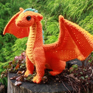 K&ouml;nnte beinhalten: Ein orangefarbener H&auml;kel-Drache mit blauen Akzenten, der auf einem Baumstumpf sitzt. Der Drache hat einen langen Hals, gro&szlig;e Fl&uuml;gel und einen kleinen Kopf mit gro&szlig;en Augen.