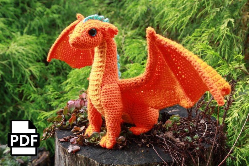 K&ouml;nnte beinhalten: Ein orangefarbener H&auml;kel-Drache mit blauen Akzenten, der auf einem Baumstumpf sitzt. Der Drache hat einen langen Hals, gro&szlig;e Fl&uuml;gel und einen kleinen Kopf mit gro&szlig;en Augen.
