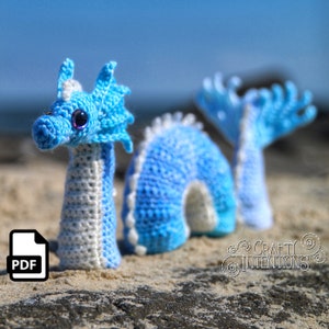 May include: A crocheted blue and white sea serpent amigurumi toy with a purple eye. The toy is sitting on a sandy beach.