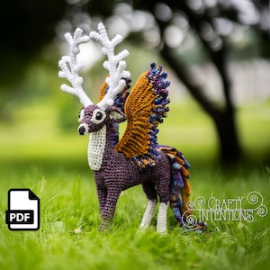 May include: A crocheted mythical creature with deer-like features, including white antlers, wings, and a purple body. The wings are a mix of orange, purple, and blue. The creature stands on green grass. A PDF icon is in the lower left corner.