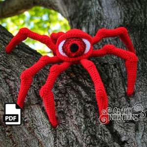 Puede incluir: Una araña roja de crochet con un ojo grande blanco y una pupila negra. La araña está sentada en un tronco de árbol.