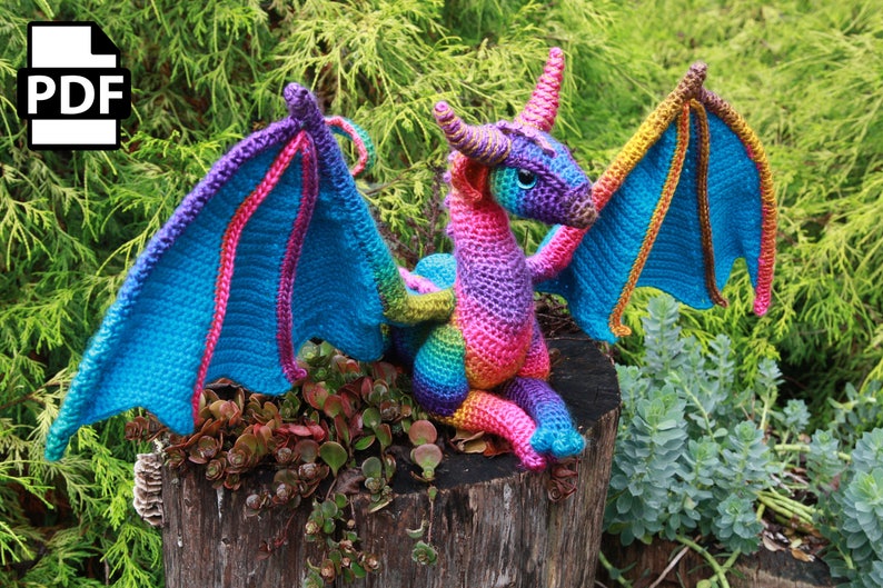May include: A colorful crocheted dragon with rainbow-colored yarn. The dragon has large wings and a long tail. It is sitting on a tree stump.