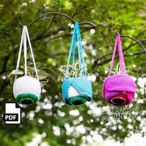 Könnte beinhalten: Drei gehäkelte Hängepflanzentopf in Weiß, Blau und Pink. Die Töpfe sind in Form von Augen gestaltet und haben eine grüne, blaue und rote Iris. Die Töpfe hängen an einem Ast.