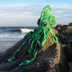 May include: Green and black crocheted squid plush toy sitting on a rock on a beach. The toy has a large head with a single eye and a long, thin body with tentacles.