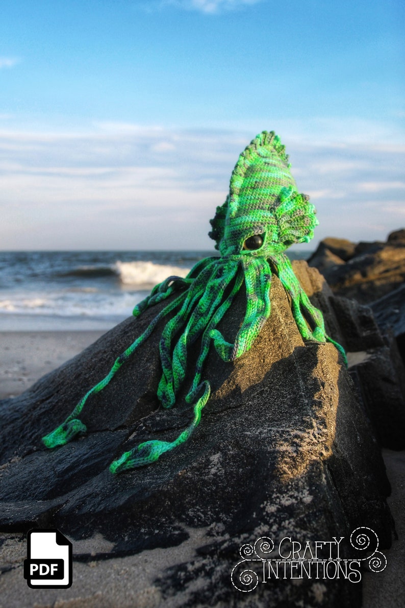 May include: Green and black crocheted squid plush toy sitting on a rock on a beach. The toy has a large head with a single eye and a long, thin body with tentacles.