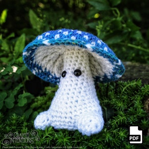May include: A crocheted mushroom amigurumi toy with a blue and white cap and a white body. The toy has black eyes and is sitting on a bed of green moss.