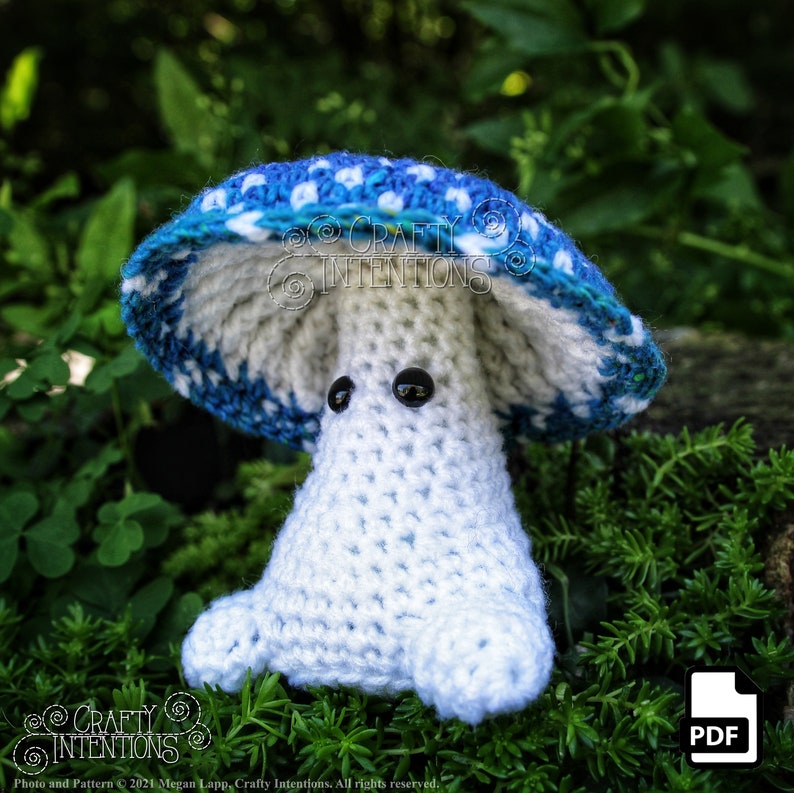 May include: A crocheted mushroom amigurumi toy with a blue and white cap and a white body. The toy has black eyes and is sitting on a bed of green moss.