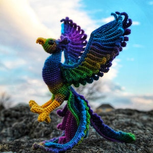 May include: A colorful crocheted phoenix with a yellow beak and legs, standing on a rocky surface. The phoenix has a purple, green, and blue color scheme. The image includes the text "CRAFTY INTENTIONS" in a circle.