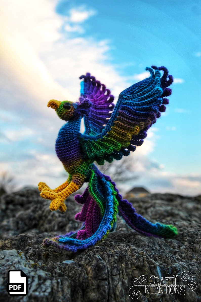 May include: A colorful crocheted phoenix with a yellow beak and legs, standing on a rocky surface. The phoenix has a purple, green, and blue color scheme. The image includes the text "CRAFTY INTENTIONS" in a circle.