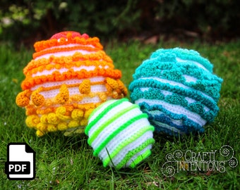 Könnte beinhalten: Drei gehäkelte Bälle in verschiedenen Farben und Mustern. Der größte Ball ist orange, gelb und weiß. Der mittlere Ball ist blau und weiß. Der kleinste Ball ist grün und weiß. Die Bälle liegen auf einem Bett aus grünem Gras.