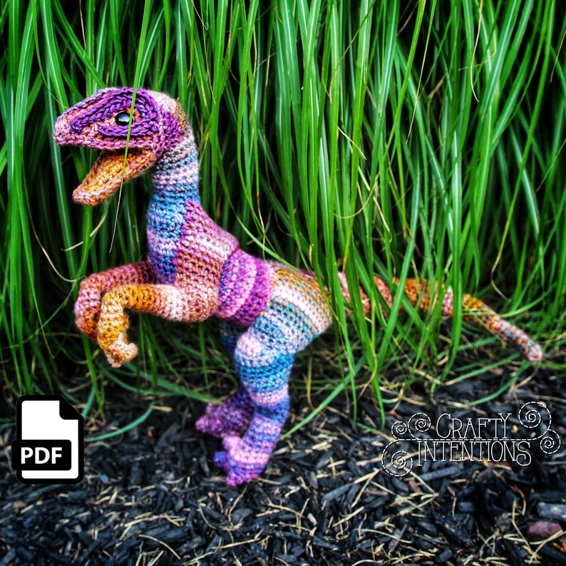May include: A colourful crocheted dinosaur toy in shades of pink, purple, blue, and orange. The dinosaur is standing on its hind legs with its tail curled around its body. The toy is sitting on a bed of brown mulch.