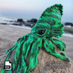 May include: A green and blue crocheted squid plush toy with a large eye. The squid is sitting on a sandy surface. The text "CRAFTY INTENTIONS" is visible in the lower right corner of the image.