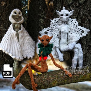 May include: Three crocheted figures, a ghost, a fairy, and a goblin, are sitting on a tree branch. The ghost is white with a hood, the fairy is white with a lacy shawl, and the goblin is brown with a green wreath around its neck. The figures are all handmade and are part of a crochet pattern.