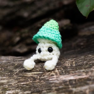 Puede incluir: Una figura de hongo de ganchillo hecha a mano con cuerpo blanco, ojos negros y gorro verde y verde claro. El hongo tiene dos peque&ntilde;os brazos y est&aacute; sobre un fondo de madera borroso. La imagen incluye el texto "Crafty Intentions" y un icono PDF.