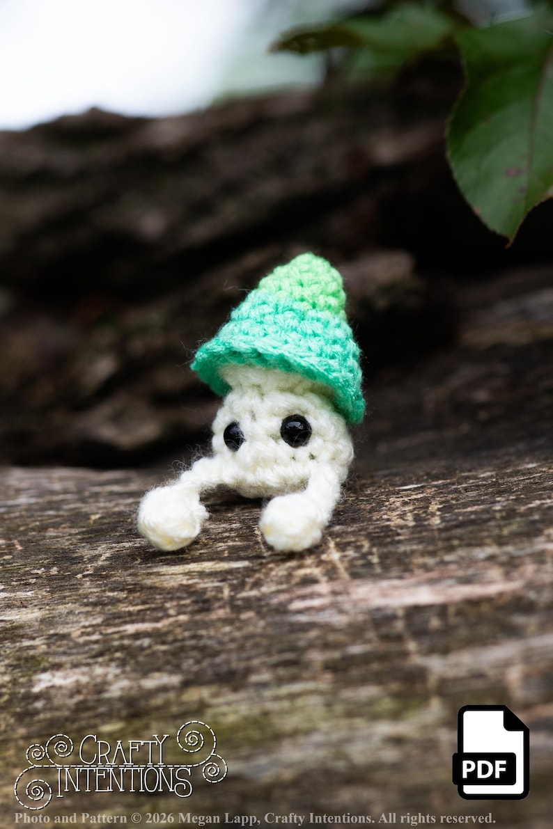 Puede incluir: Una figura de hongo de ganchillo hecha a mano con cuerpo blanco, ojos negros y gorro verde y verde claro. El hongo tiene dos peque&ntilde;os brazos y est&aacute; sobre un fondo de madera borroso. La imagen incluye el texto "Crafty Intentions" y un icono PDF.