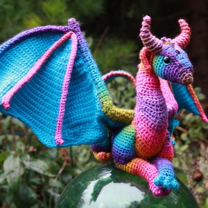 K&ouml;nnte beinhalten: Ein bunter H&auml;kel-Drache mit einem Regenbogenmuster. Der Drache hat einen blauen, rosa und violetten K&ouml;rper mit gr&uuml;nen Fl&uuml;geln. Der Drache sitzt auf einer gr&uuml;nen Glasfl&auml;che.