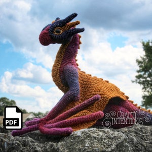 May include: A crocheted dragon amigurumi toy with a purple, orange, and brown color scheme. The dragon is sitting on a rock with a cloudy sky in the background.