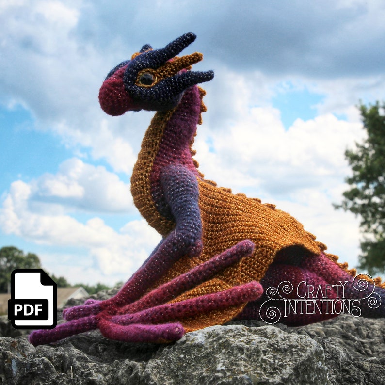 May include: A crocheted dragon amigurumi toy with a purple, orange, and brown color scheme. The dragon is sitting on a rock with a cloudy sky in the background.