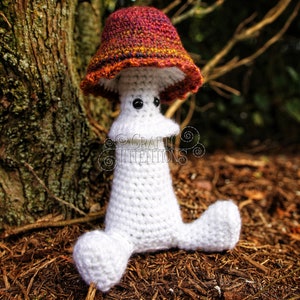 May include: A white crocheted mushroom amigurumi toy with a red, orange, and yellow crocheted cap. The toy is sitting on a bed of brown pine needles. The text "CRAFTY INTENTIONS" is visible on the toy.