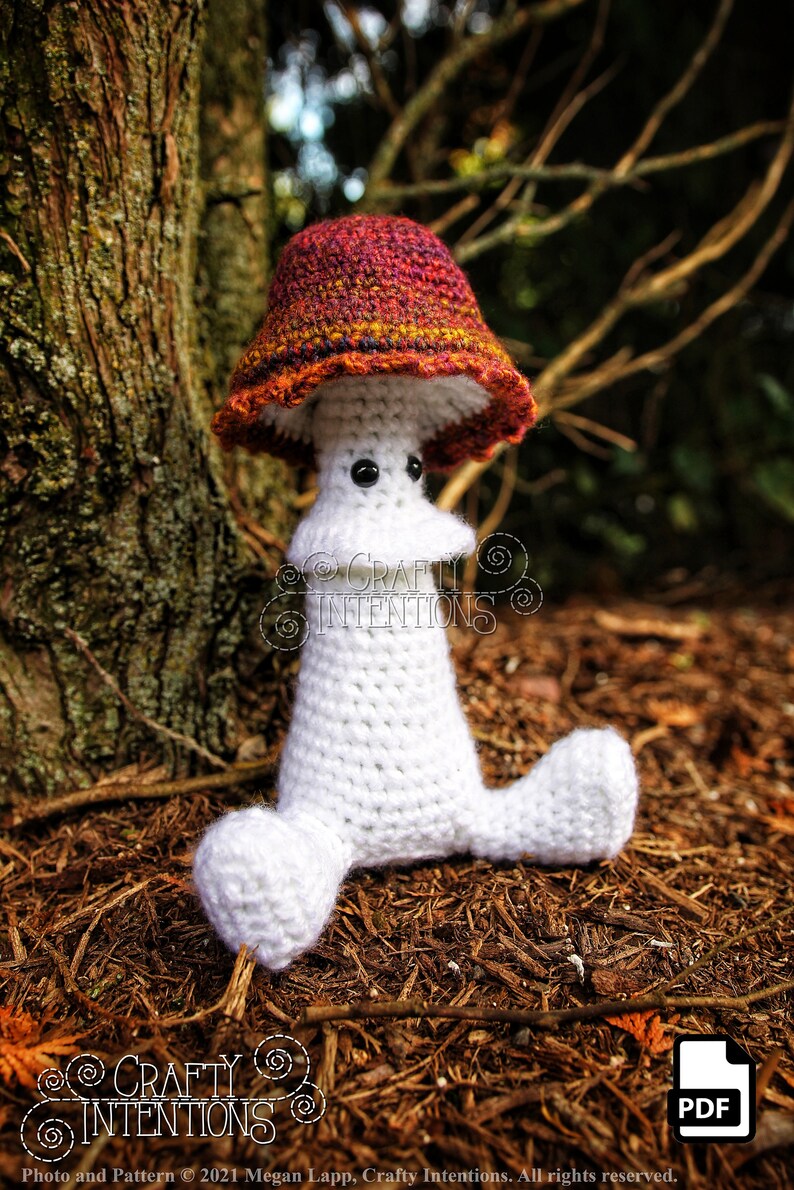 May include: A white crocheted mushroom amigurumi toy with a red, orange, and yellow crocheted cap. The toy is sitting on a bed of brown pine needles. The text "CRAFTY INTENTIONS" is visible on the toy.