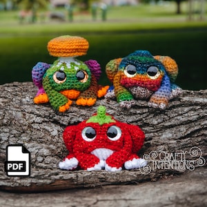 Może przedstawiać: Trzy pluszowe zabawki amigurumi: czerwona papryka, zielono-pomarańczowy grzyb i tęczowa żaba. Zabawki leżą na pniu drzewa. Obrazek zawiera tekst "CRAFTY INTENTIONS".