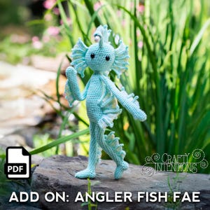 May include: A light blue crocheted Angler Fish Fae doll stands on a rock in front of greenery. The doll has large, detailed fins and a whimsical design. The text "ADD ON: ANGLER FISH FAE" is visible at the bottom of the image, along with a PDF icon.