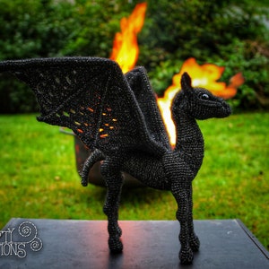 May include: A black crocheted dragon with wings, standing on a black surface in front of a fire. The dragon has a long neck and a small head with a pointed snout. The wings are spread out and the dragon is looking to the right. The fire is behind the dragon and is burning brightly. The background is a green lawn.