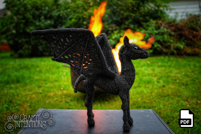 May include: A black crocheted dragon with wings, standing on a black surface in front of a fire. The dragon has a long neck and a small head with a pointed snout. The wings are spread out and the dragon is looking to the right. The fire is behind the dragon and is burning brightly. The background is a green lawn.