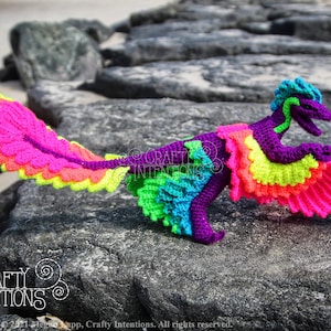 May include: A colourful crocheted dinosaur with a rainbow tail and wings. The dinosaur is purple with green accents and is posed on a rock.