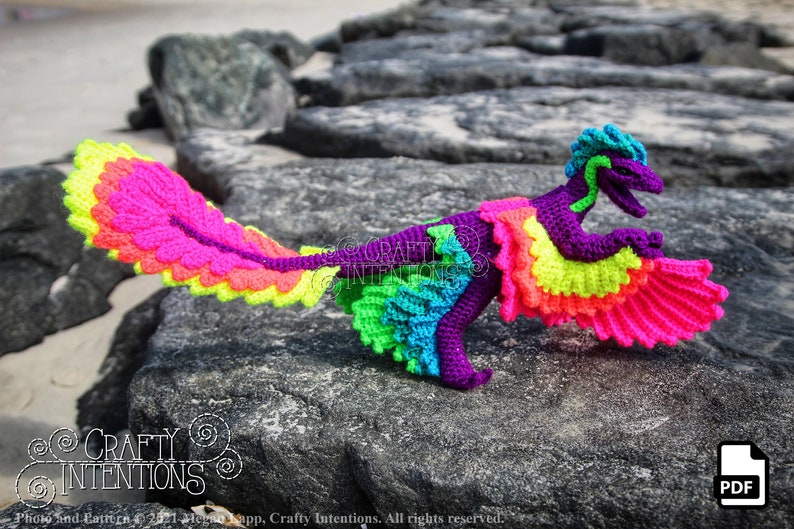 May include: A colourful crocheted dinosaur with a rainbow tail and wings. The dinosaur is purple with green accents and is posed on a rock.