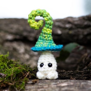 Puede incluir: Un fantasma de ganchillo hecho a mano con un sombrero de bruja verde y azul. El fantasma es blanco con ojos negros y pies peque&ntilde;os. El sombrero tiene una parte superior rizada. El fondo es un entorno natural borroso. La imagen incluye el texto "Crafty Intentions" y un icono PDF.