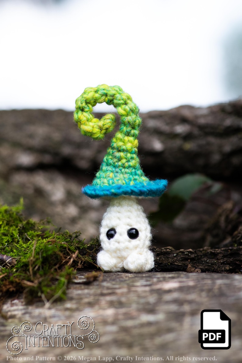 Puede incluir: Un fantasma de ganchillo hecho a mano con un sombrero de bruja verde y azul. El fantasma es blanco con ojos negros y pies peque&ntilde;os. El sombrero tiene una parte superior rizada. El fondo es un entorno natural borroso. La imagen incluye el texto "Crafty Intentions" y un icono PDF.