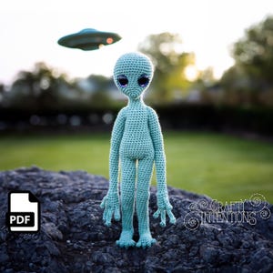 May include: A crocheted alien doll with large blue eyes standing on a rock with a flying saucer in the background. The doll is light green and has long arms and legs. The doll is wearing a simple, crocheted outfit. The background is a blurry image of a green field and trees.