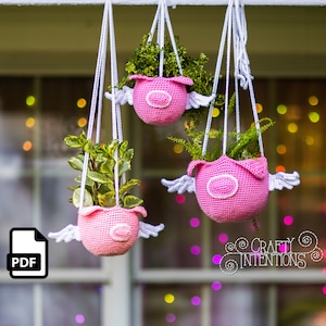 May include: Three pink crocheted hanging planters shaped like pigs with white wings. Each planter has a small green plant inside. The planters are hanging from a white string.