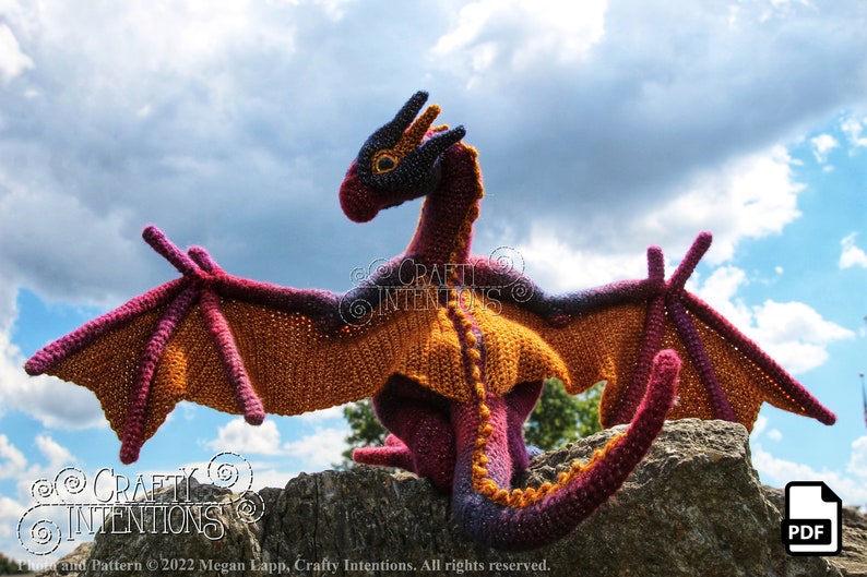 May include: A crocheted dragon with purple, orange, and black yarn. The dragon is sitting on a rock with its wings spread out. The background is a blue sky with white clouds.