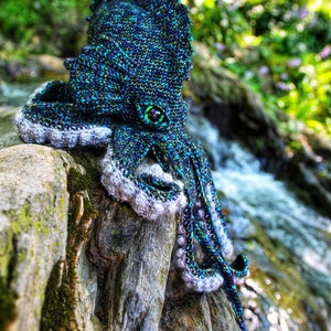 May include: A crocheted octopus with a blue, green, and black color scheme. The octopus is sitting on a mossy log with a small stream in the background. The octopus has a large, round eye with a green iris.