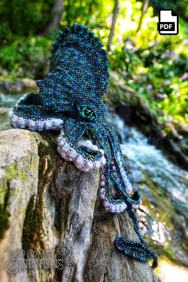 May include: A crocheted octopus with a blue, green, and black color scheme. The octopus is sitting on a mossy log with a small stream in the background. The octopus has a large, round eye with a green iris.