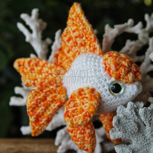 Puede incluir: Un juguete amigurumi de pez dorado tejido a crochet con hilo naranja y blanco. El juguete tiene un cuerpo blanco y aletas y cola naranja. El juguete est&aacute; sentado sobre una superficie blanca similar al coral.