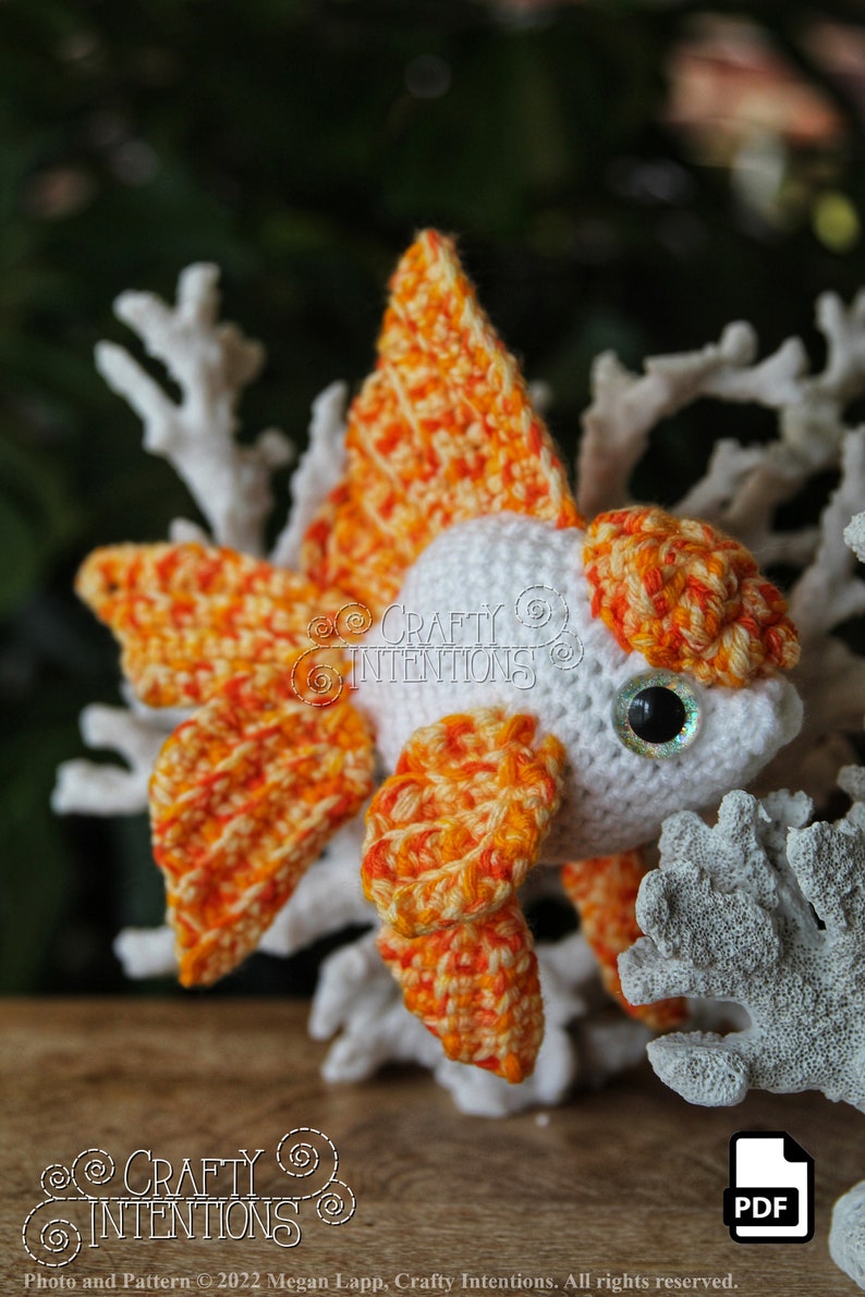 Puede incluir: Un juguete amigurumi de pez dorado tejido a crochet con hilo naranja y blanco. El juguete tiene un cuerpo blanco y aletas y cola naranja. El juguete est&aacute; sentado sobre una superficie blanca similar al coral.