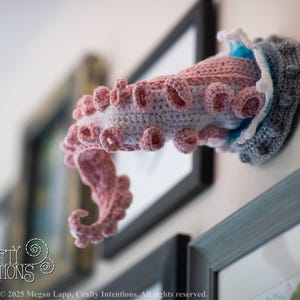 May include: A crocheted octopus tentacle wall hanging in shades of pink, gray, and blue. The handmade art piece is mounted on a wall, with a PDF icon in the lower right corner. The Crafty Intentions logo is visible in the lower left corner.