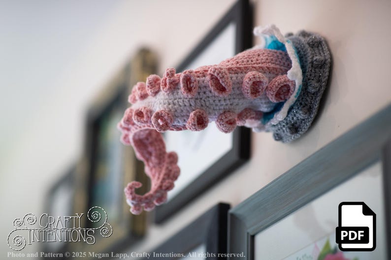 May include: A crocheted octopus tentacle wall hanging in shades of pink, gray, and blue. The handmade art piece is mounted on a wall, with a PDF icon in the lower right corner. The Crafty Intentions logo is visible in the lower left corner.