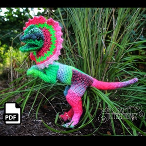 May include: A crocheted dinosaur toy with a pink, green and blue colour scheme. The dinosaur is standing on a bed of green grass. The toy is available as a PDF download.