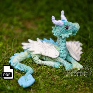 May include: A crocheted dragon in shades of blue, green, and white, resting on green grass. The dragon has white wings, blue horns, and a detailed, textured body. The dragon's eyes are black. The image includes a PDF icon and the text "Crafty Intentions".