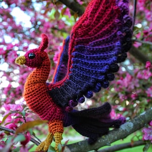 May include: A crocheted phoenix with red, orange, purple, and black yarn. The phoenix is perched on a branch with pink blossoms. The image includes the text "CRAFTY INTENTIONS" in a circle.