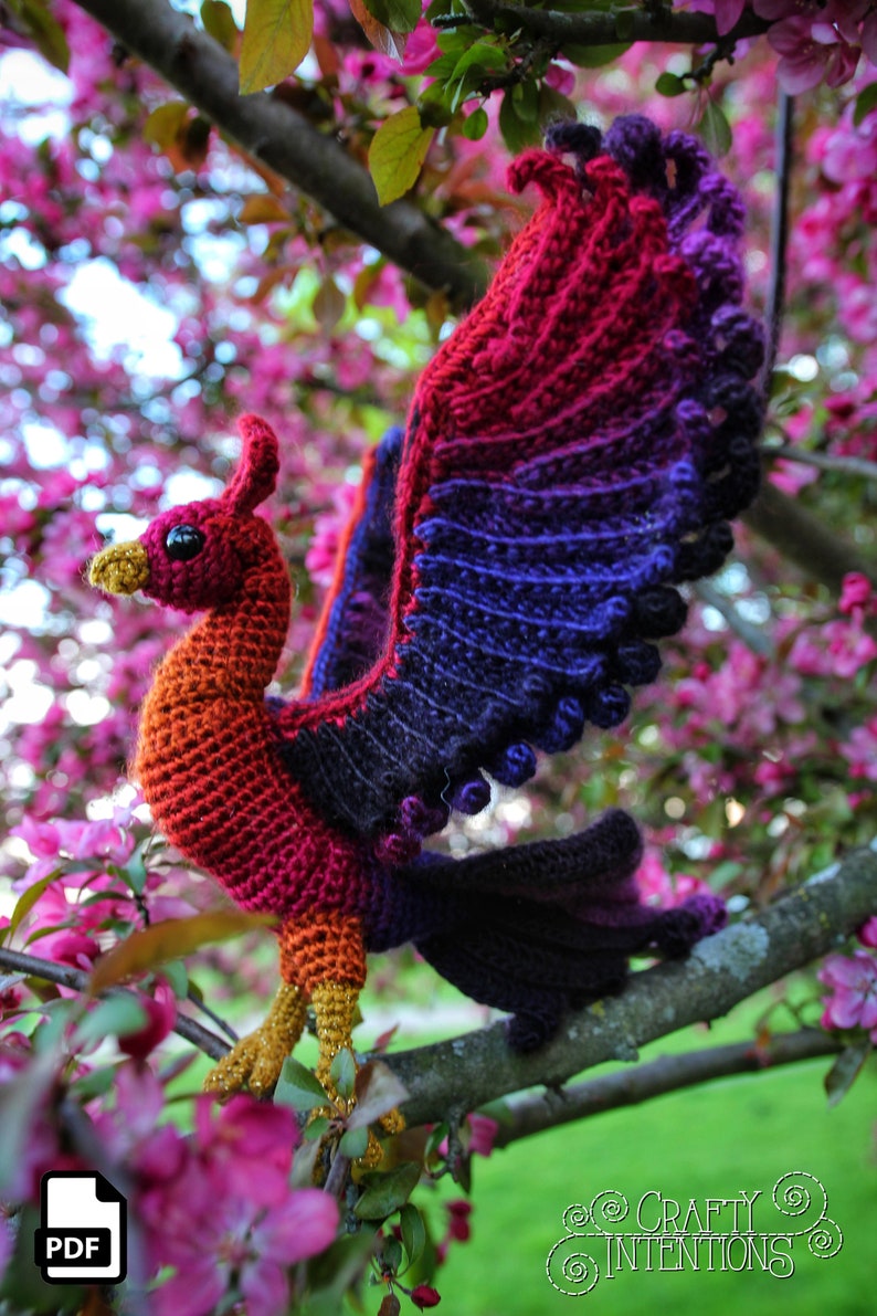 May include: A crocheted phoenix with red, orange, purple, and black yarn. The phoenix is perched on a branch with pink blossoms. The image includes the text "CRAFTY INTENTIONS" in a circle.
