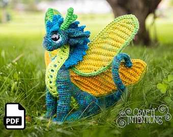 May include: A handmade crochet dragon, featuring blue, green, and yellow hues, with prominent wings and intricate detailing. The dragon is positioned in a grassy outdoor environment. A PDF icon is visible in the bottom left.