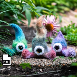 Peut inclure: Trois créatures amigurumi crochetées avec de grands yeux et du fil coloré. Les créatures sont assises sur un lit de gravier et de feuillage vert. Les créatures sont bleu sarcelle et vert, arc-en-ciel et violet et rose.